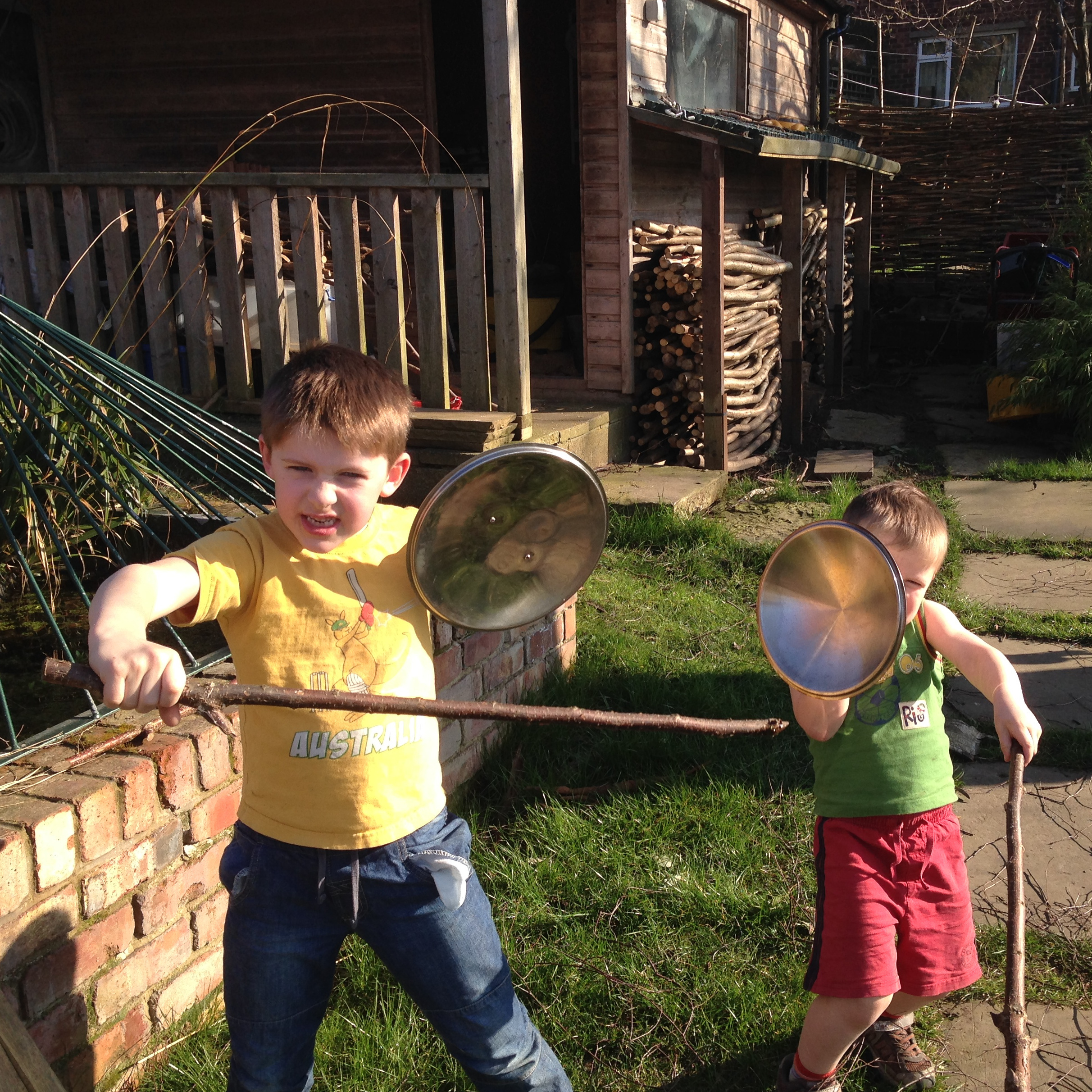 2 boys holding sticks and saucepan lids, pretending to fight