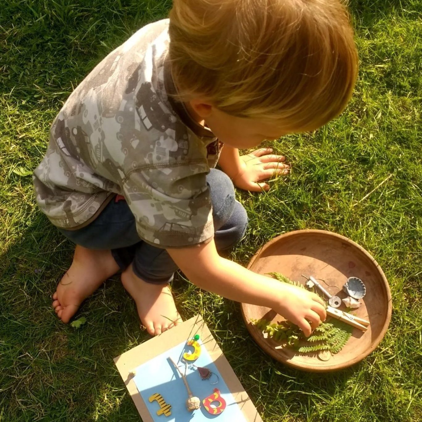 small child kneeling on grass with their hand in a bowl of ferns and shells and other natural objects