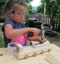 girl wearing safety goggles hammering a nail into a wooden train that she is making