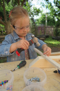 little girl in denim jacket and plastic goggles, hammering a nail into a piece of wood, on a workbench with pots of nails and screws and a screwdriver