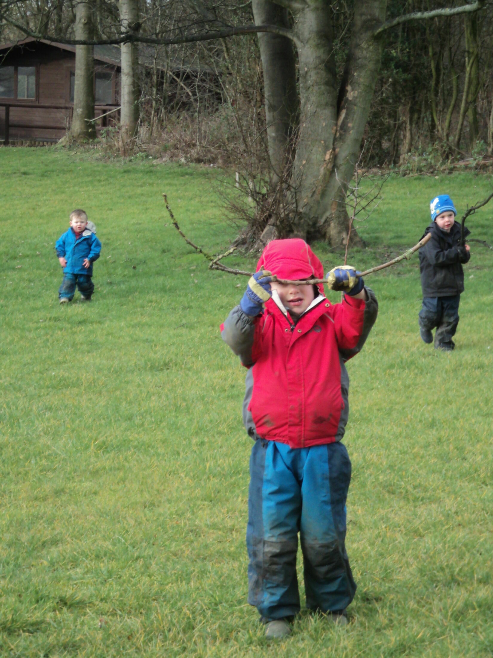 boy in red coat and blue waterproof trousers in foreground, holding up a long stick, with 2 small children also in outdoor gear, running behind