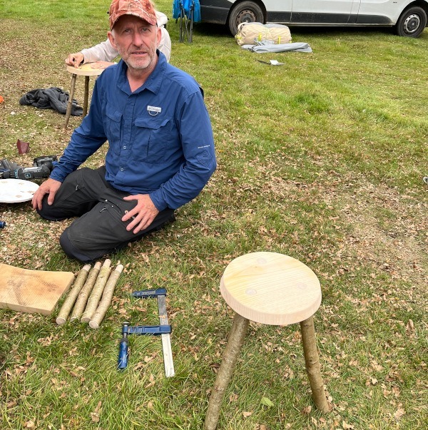 a man in a blue shirt and grey jeans kneeling on the grass nexdt to some wooden sticks and a saw, with a small handmade stool