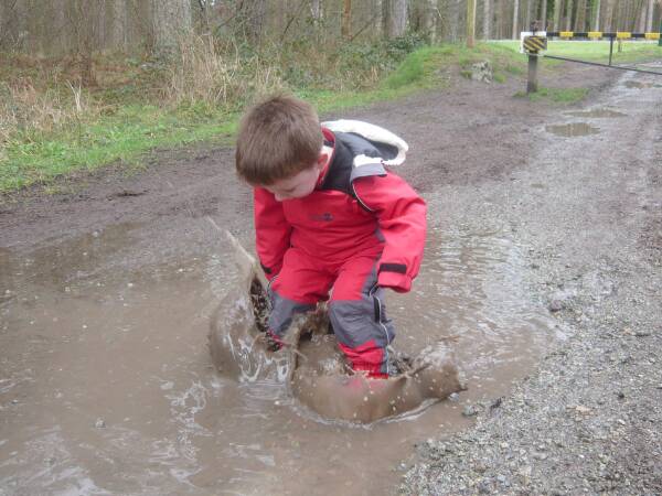 Nursery aged child in waterproof clothing and wellies splashing in a muddy puddle.